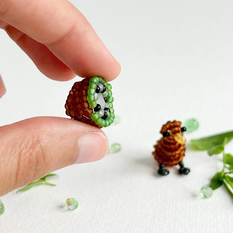 K&ouml;nnte beinhalten: Ein geperltes Kiwi-Frucht und ein geperlter Kiwi-Vogel, beide braun und gr&uuml;n, sind auf einer wei&szlig;en Oberfl&auml;che zu sehen. Die Kiwi-Frucht wird von einer Hand gehalten.