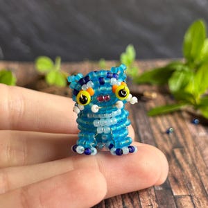 May include: A small, beaded creature figurine in shades of blue, white, and yellow. The handmade figure has large yellow eyes with black pupils, a red mouth, and small white accents. The figurine is standing upright.