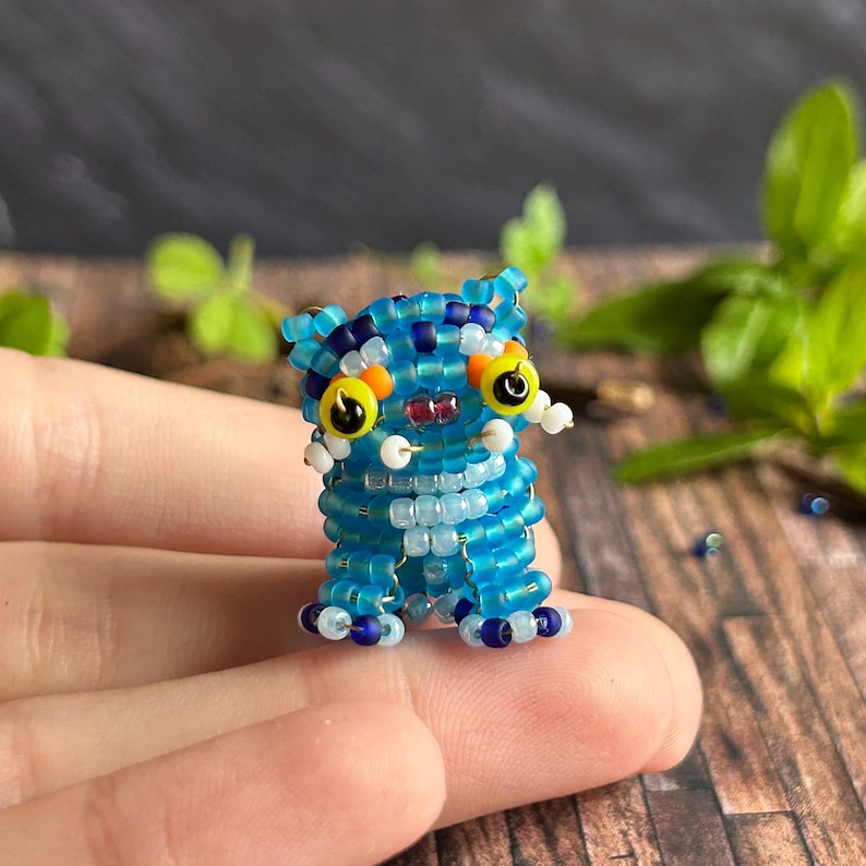 May include: A small, beaded creature figurine in shades of blue, white, and yellow. The handmade figure has large yellow eyes with black pupils, a red mouth, and small white accents. The figurine is standing upright.