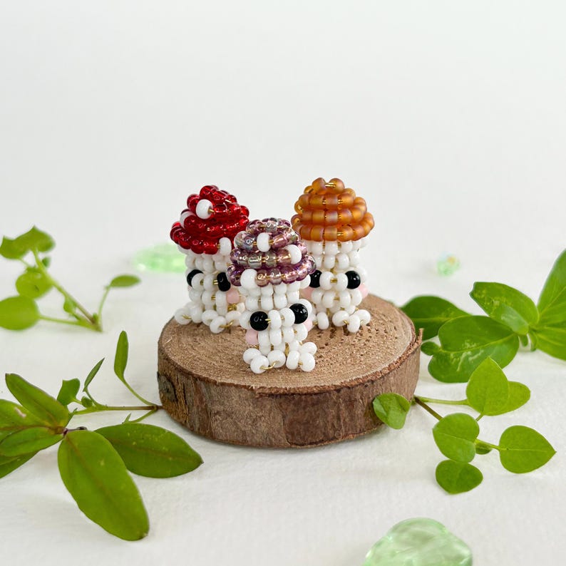 May include: Three small beaded mushroom figurines with different colored caps. The mushrooms are white with black eyes and pink cheeks. They are sitting on a small wooden slice.