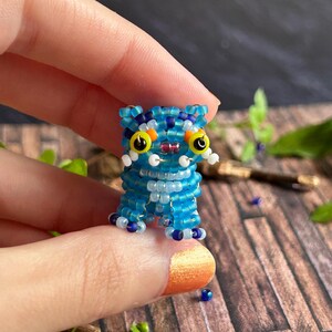 May include: A miniature beaded cat figure, predominantly blue with white, yellow, and purple details. The cat has large yellow eyes and small white whiskers. The figure is held between fingers.
