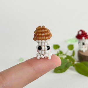 May include: A small, white beaded mushroom with a brown cap and black eyes. The mushroom is sitting on a finger.