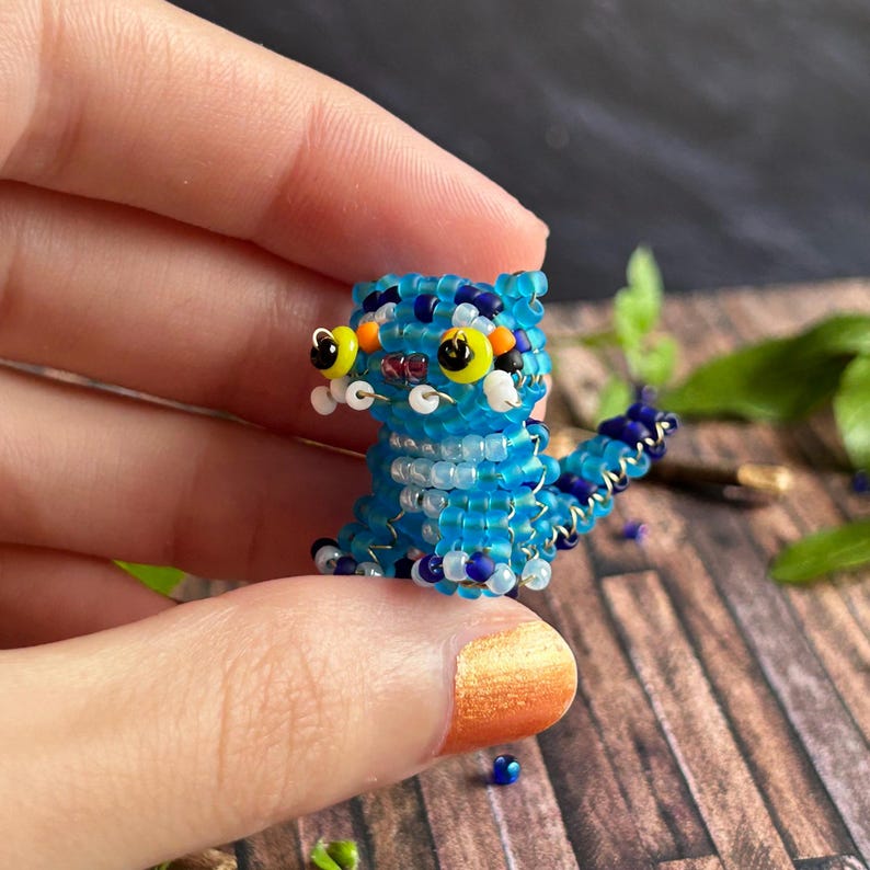 May include: A small, beaded creature figurine in shades of blue, white, and navy. The figurine has large yellow eyes with black pupils, and white accents. The tail is a darker blue with a black tip. The figurine is held in a hand.
