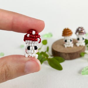 May include: A close-up of a small, red and white beaded mushroom with a black bead for an eye. The mushroom is being held by a finger. There are two other mushrooms in the background, one brown and one purple.