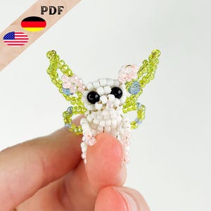 Könnte beinhalten: Eine weiße Perlenfee mit grünen und rosa Flügeln. Die Fee hat schwarze Augen und einen weißen Körper. Die Fee wird von einer Hand gehalten.