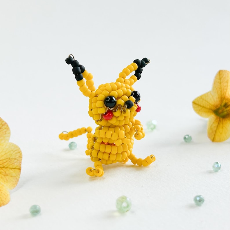 May include: A handmade yellow beaded Pikachu figure with black ears and eyes, and red cheeks. The figure is posed with arms outstretched and legs bent, giving it a playful appearance. The figure is surrounded by small, light green beads and yellow flowers.