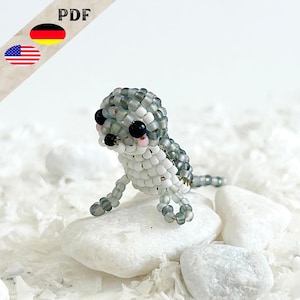 Może przedstawiać: Mała, szara i biała figurka zwierzęcia zrobiona z koralików z czarnymi oczami i różowymi policzkami. Figurka siedzi na białym kamieniu.