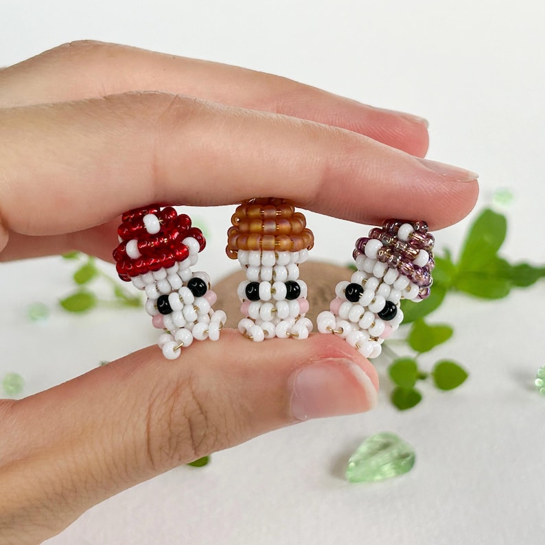 May include: Three beaded mushroom figurines with different colored caps. The mushrooms are white with pink cheeks and black eyes. The caps are red, brown, and purple.