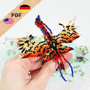 Op de afbeelding: Een kralen vlinder in oranje, zwart en geel, met blauwe en zilveren accenten. De vleugels zijn gedetailleerd. De afbeelding bevat de tekst "PDF" en de vlaggen van de VS en Duitsland.