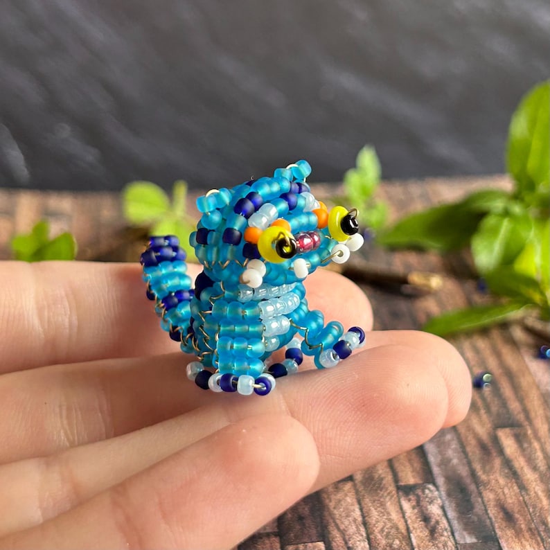 May include: A small, beaded creature in shades of blue, white, and yellow. The handmade figurine has large, round eyes and a detailed, textured body. The creature is sitting, with its legs and tail curled.
