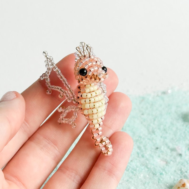 May include: A beaded seahorse figurine in shades of cream, peach, and clear. The seahorse has black eyes and a detailed, textured body. The wings and crown are made of clear beads. The figurine is held in a hand against a light blue background.