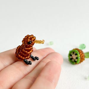 K&ouml;nnte beinhalten: Eine kleine braune Perlenfigur eines Kiwi-Vogels mit schwarzen Augen und einem gelben Schnabel. Die Figur sitzt auf einer Hand.