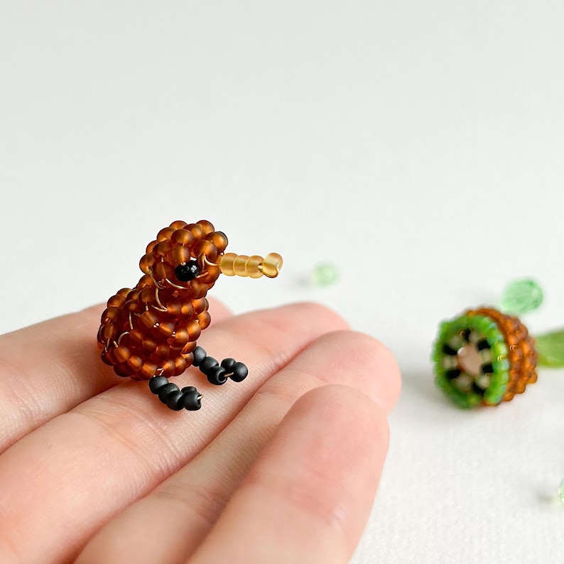K&ouml;nnte beinhalten: Eine kleine braune Perlenfigur eines Kiwi-Vogels mit schwarzen Augen und einem gelben Schnabel. Die Figur sitzt auf einer Hand.