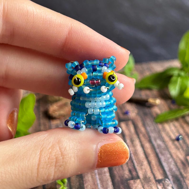 May include: A small, beaded cat figurine in shades of blue, white, and dark blue. The cat has large yellow eyes with black pupils, white whiskers, and a small orange nose. The figurine is made of small beads.