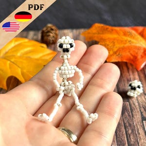 Peut inclure: Une petite figurine de squelette en perles blanches avec des yeux noirs est posée sur une main. Le squelette est assis les jambes croisées et les bras tendus.