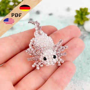Könnte beinhalten: Eine kleine, dreidimensionale Axolotl-Figur, gefertigt aus winzigen, durchscheinenden weißen und blassrosa Glasperlen. Das Wesen hat einen abgerundeten Körper, einen langen Schwanz, vier kleine Beine und äußere Kiemen, akzentuiert durch zwei schwarze Perlen.