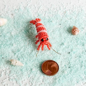 May include: A handcrafted beaded shrimp figurine in red and white, with black eyes and wire antennae. The shrimp is placed on a bed of light blue sand, next to a one-cent coin and seashells. The shrimp measures approximately 1 inch in length.