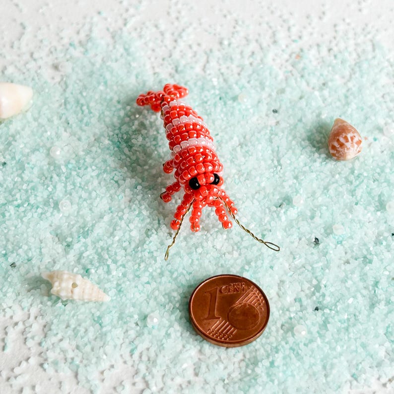 May include: A handcrafted beaded shrimp figurine in red and white, with black eyes and wire antennae. The shrimp is placed on a bed of light blue sand, next to a one-cent coin and seashells. The shrimp measures approximately 1 inch in length.