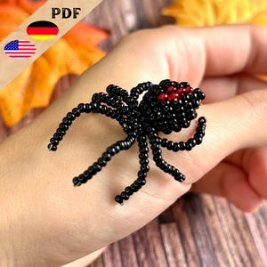 Può includere: Ragno nero di perline con accenti di perline rosse. Il ragno è lungo circa 5 cm ed è fatto di perline nere e rosse. Il ragno è sulla mano di una persona.