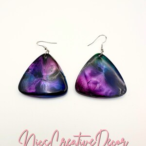 Puede incluir: Un par de pendientes de forma triangular con un diseño abstracto en morado, azul y verde. Los pendientes tienen ganchos plateados.