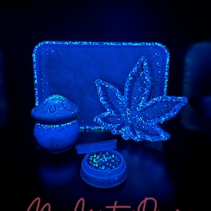 May include: A collection of blue resin accessories, including a tray, a mushroom-shaped container, a cannabis leaf, and a grinder. The items are illuminated with a blue light, and the surfaces are speckled with glitter.