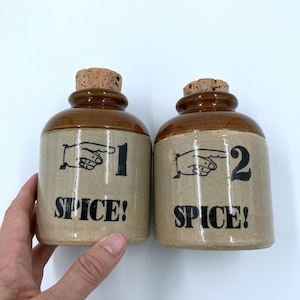 Puede incluir: Dos tarros de especias de cerámica marrón con tapas de corcho. Los tarros están etiquetados con "1 SPICE!" y "2 SPICE!" con una mano apuntando al número.
