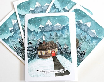 May include: A set of five greeting cards featuring a watercolor illustration of a cozy cabin in a snowy forest with mountains in the background. The text on the card reads "Sending you peace and light".