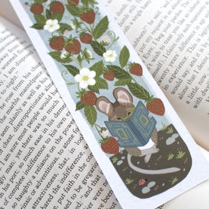 May include: A whimsical bookmark featuring a mouse reading a book amidst a strawberry patch. The illustration includes ripe red strawberries, white flowers, and green leaves, all set against a light blue background. The mouse is wearing glasses.