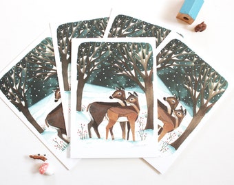Pode incluir: Um conjunto de cartões de felicitações retangulares com uma ilustração em aguarela de dois veados castanhos num bosque nevado com árvores escuras e flocos de neve brancos. A cena inclui pequenas bagas vermelhas em ramos finos.