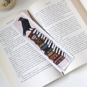 May include: A bookmark featuring a black raven perched on a stack of vintage books. The raven is looking to the right, and the books are stacked in a staggered pattern. The background is a soft pink.