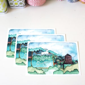 May include: Three watercolor postcards featuring a stylized illustration of the state of Oregon with the text "Portland Oregon" and a deer jumping over the state border. The top postcard also features a water tower with the text "Old Town".