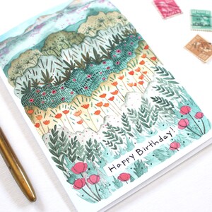 May include: Watercolour birthday card depicting a layered landscape with green, orange, and pink floral elements. The card reads "Happy Birthday!" and is next to a brass pen and vintage stamps.