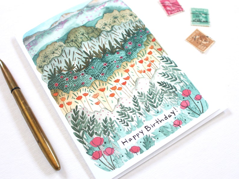 May include: Watercolour birthday card depicting a layered landscape with green, orange, and pink floral elements. The card reads "Happy Birthday!" and is next to a brass pen and vintage stamps.