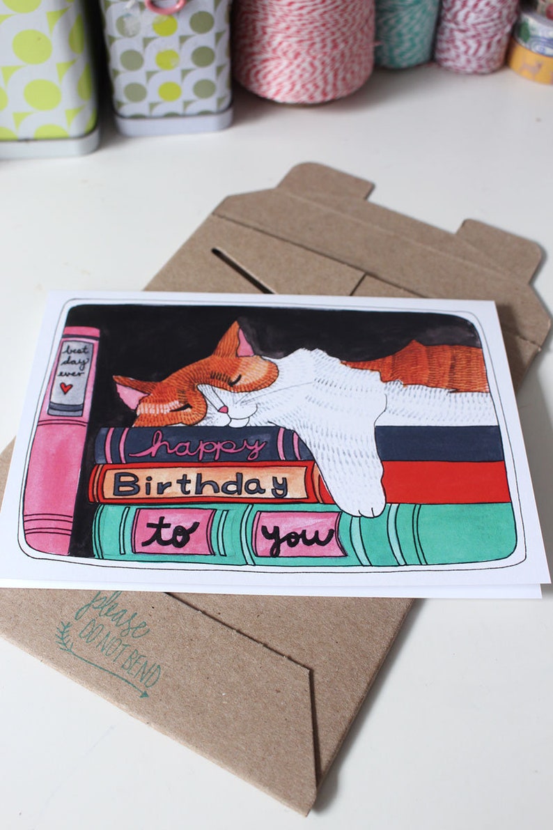 K&ouml;nnte beinhalten: Eine Geburtstagskarte mit einer Katze, die auf einem Stapel bunter B&uuml;cher schl&auml;ft. Die Karte lautet "Happy Birthday to you".