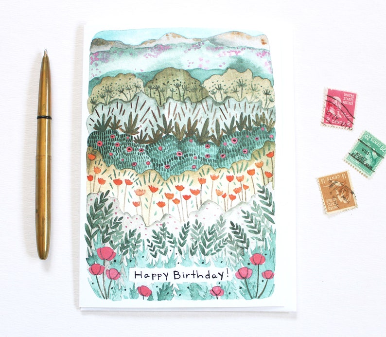 May include: A watercolour birthday card depicting a layered landscape with green, orange, and pink floral elements. The card includes the text "Happy Birthday!" and is accompanied by a gold pen and three vintage postage stamps.
