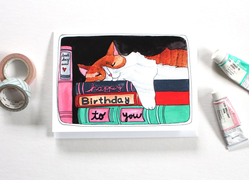 K&ouml;nnte beinhalten: Eine Gru&szlig;karte mit einer Illustration einer orange-wei&szlig;en Katze, die auf einem Stapel B&uuml;cher schl&auml;ft. Die B&uuml;cher haben Titel wie "bester Tag aller Zeiten" und "alles Gute zum Geburtstag an dich".