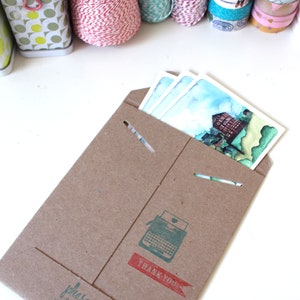May include: A brown paper envelope with a typewriter graphic and the text "Thank You" in red. The envelope is open and contains three watercolor postcards with a blue sky and a building with the text "Old Town" on it.