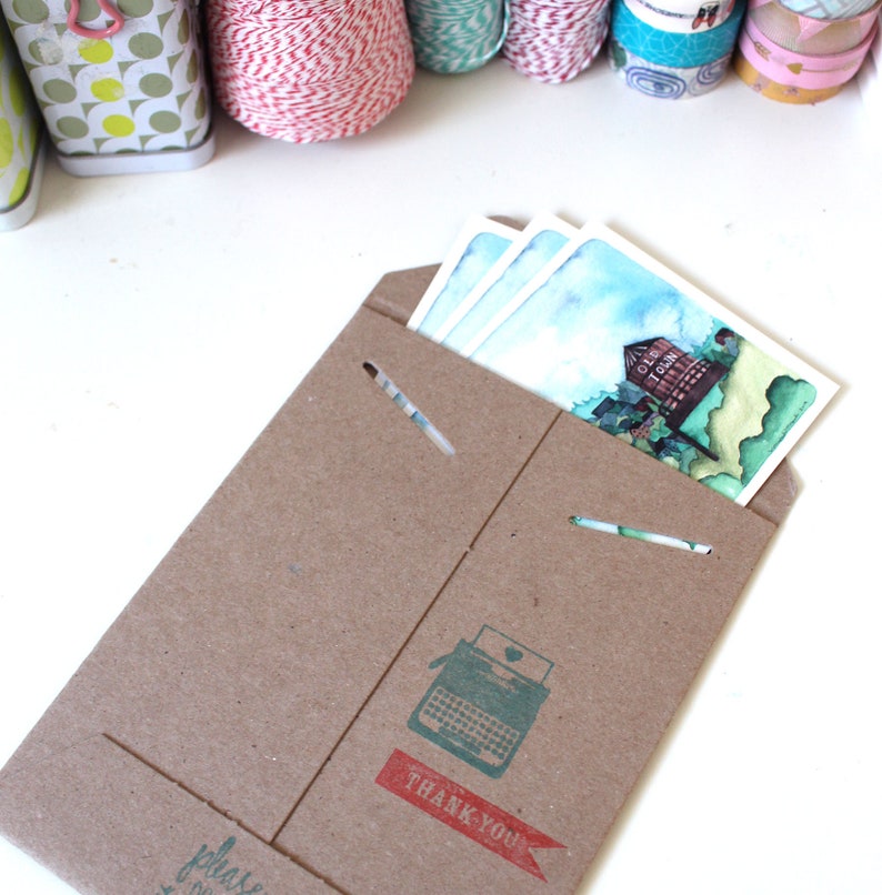 May include: A brown paper envelope with a typewriter graphic and the text "Thank You" in red. The envelope is open and contains three watercolor postcards with a blue sky and a building with the text "Old Town" on it.