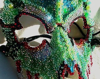 May include: A green, red, and black masquerade mask made of iridescent beads. The mask is shaped like a dragon's head with a sharp snout and pointy teeth.