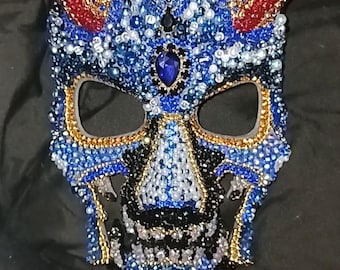 May include: A blue and gold beaded mask with a skull design. The mask has a large, blue gemstone in the center of the forehead and two horns on the top.