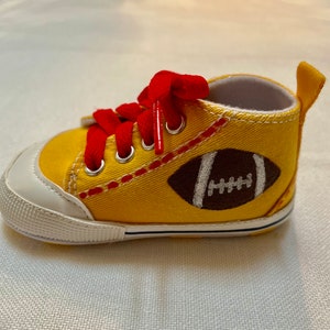 May include: Yellow canvas baby shoes with red laces and a brown football design. The shoes have white stitching and a white sole.