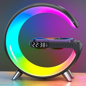 May include: A black, G-shaped lamp with a color-changing LED light. The lamp has a built-in wireless charger and a digital clock displaying 22:38. A smartphone rests on the charging pad. The lamp is supported by three silver legs.