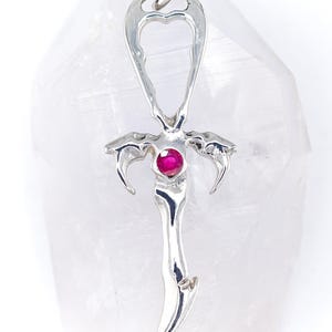 May include: A silver pendant with a heart-shaped top and a dagger-like design. The pendant features a small, round, red gemstone at its center and curved, claw-like elements. The overall design is intricate and detailed.