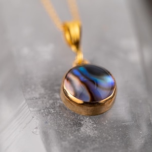 May include: A gold pendant necklace featuring a round, iridescent abalone shell. The shell displays a mix of blue, purple, and black hues, set in a gold bezel. The chain is also gold, creating a classic jewelry piece.
