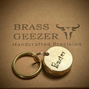 May include: Brass key ring with a round tag that says "Baxter" on it. The keyring is on a brown surface with the text "BRASS GEEZER Handcrafted Precision" and a bull's head logo.