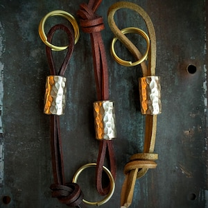 Könnte beinhalten: Drei Leder-Schlüsselanhänger mit gehämmerten Metallakzenten. Jeder Schlüsselanhänger hat einen Messingring und ein Lederband in einer anderen Farbe: Braun, Dunkelbraun und Beige.