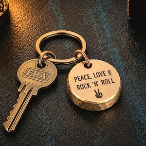 May include: A brass keychain featuring a round, hammered-texture pendant engraved with the text PEACE, LOVE AND ROCK 'N' ROLL and a peace sign symbol, attached to a split ring alongside a vintage-style key with the word ERA embossed on its head.