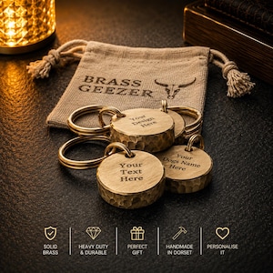 May include: Three round brass keychains with hammered edges and split rings, resting in front of a tan drawstring pouch printed with the text BRASS GEEZER and a bull icon. The keychains feature engraved text reading Your Design Here, Your Text Here, and Your Dogs Name Here.
