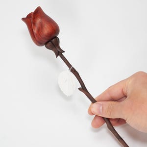 May include: A handcrafted wooden rose with a deep reddish-brown bloom and stem. The rose is held against a white background. A small, white, leaf-shaped element is attached to the stem. The rose is a decorative item.
