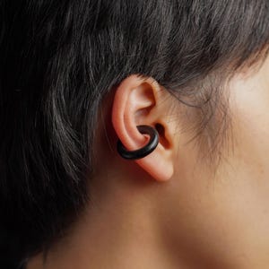 May include: A smooth, matte black circular ear cuff worn on the outer cartilage of the ear. The jewellery piece features a simple, continuous ring design with a solid, dark finish that contrasts against the skin of the ear.